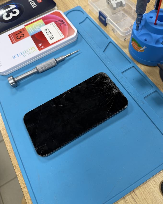 iPhone 13 con lo schermo rotto sul tappetino da riparazione, accanto a un cacciavite e alla scatola del display di ricambio